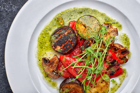 Grilled baked vegetables, eggplant, zucchini, bell pepper, champignon mushrooms, on a white plate against a dark background, with microgreenの写真素材
