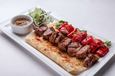 Meat shish kebab on pita bread with marinated onion sauce and baked bell pepper on a white plate on a white plateの写真素材