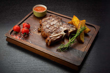 T-bone steak on a wooden board with potatoes, sauce, cherry pickers, on a dark tableの写真素材