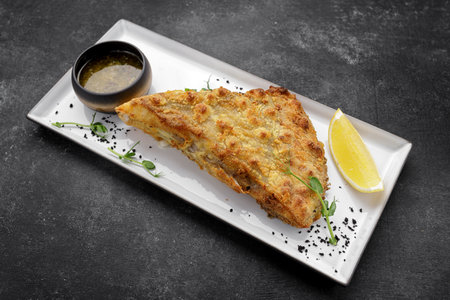 A piece of fried flounder fish with lemon on a plate, on a dark background