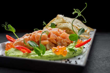 Salmon tartar with red, avocado puree, caviar and croutons. On a plate, on a dark backgroundの写真素材