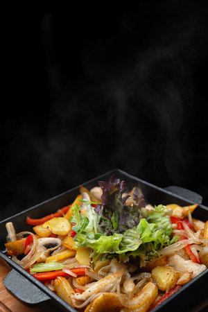Fried stewed potatoes with vegetables and mushrooms in a cast-iron skillet in a wooden pan, on a black backgroundの写真素材