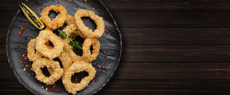 Fried onion rings in batter with spices, on a black plate, on a wooden background. Banner