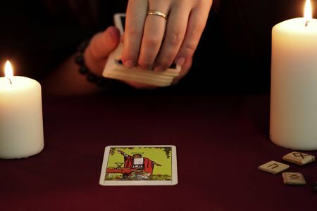 A witch is fortune teller in a black mantle pulls a tarot card from deck. Tarot cards, ancient runes and white candles. Occult, esoteric, divination and wicca concept. Mystic and vintage background.のeditorial素材
