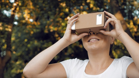 Woman explores virtual reality using cardboard VR glasses. Augmented reality quickly comes in human lives. VR mounted displays helmets and glasses are the most popular devices for playing AR games.の写真素材