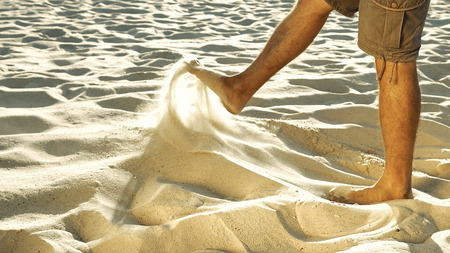 Legs of tanned man in khaki shorts sieving sand with feet. Barefoot human playing in desert. Senseless act and loneliness concept. Close-up. Side view.の写真素材