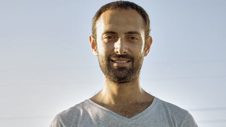 Tanned man with beard and in gray t-shirt smiles with teeth. Cheerful person. Human under sunlight. Joy, happiness and consent concept. Close-up. Front view.の写真素材
