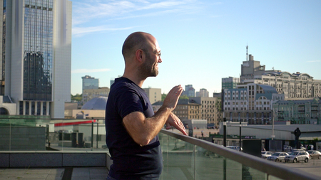 Handsome man is standing on the balcony terrace in the urban city centre. Cheerful man in glasses at sunset against the blue sky at city downtown.の写真素材
