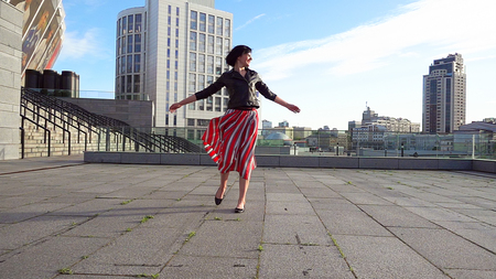 Beautiful brunette woman is dancing feeling happy outdoors. City life urban young happy people behavior. Sunset cityscape clear sky shot. The woman wears long striped pleated skirt.の写真素材