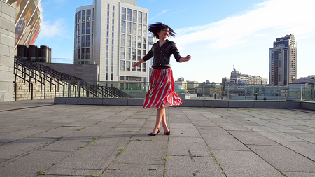 Beautiful brunette woman is dancing feeling happy outdoors. City life urban young happy people behavior. Sunset cityscape clear sky shot. The woman wears long striped pleated skirt.の写真素材