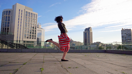 Beautiful brunette woman is dancing feeling happy outdoors. City life urban young happy people behavior. Sunset cityscape clear sky shot. The woman wears long striped pleated skirt.の写真素材