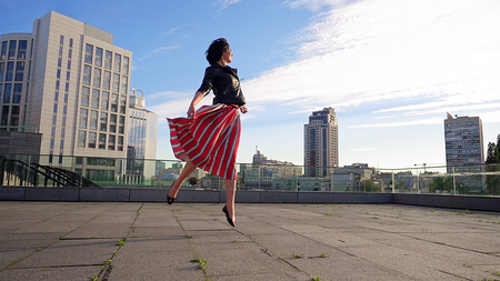 Beautiful brunette woman is dancing feeling happy outdoors. City life urban young happy people behavior. Sunset cityscape clear sky shot. The woman wears long striped pleated skirt.の写真素材