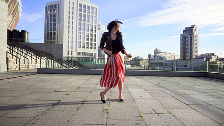 Beautiful brunette woman is dancing feeling happy outdoors. City life urban young happy people behavior. Sunset cityscape clear sky shot. The woman wears long striped pleated skirt.の写真素材