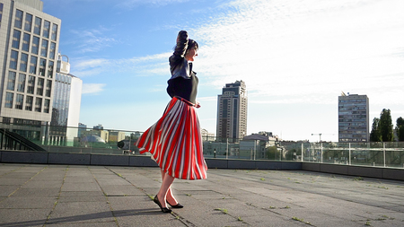 Beautiful brunette woman is dancing feeling happy outdoors. City life urban young happy people behavior. Sunset cityscape clear sky shot. The woman wears long striped pleated skirt.の写真素材