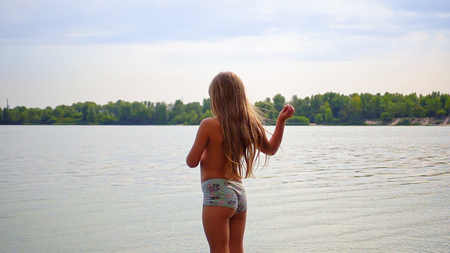Little cute blond longhair girl is standing at riverside bank coast. Family recreational activity outdoors. Sunset shot.の写真素材