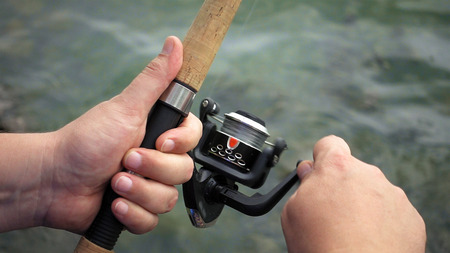 Fishing reel closeup. Man is fishing with spinning fishing rod close to coast shore line. Outdoor recreational activity.の写真素材