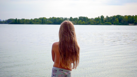 Little cute blond longhair girl is standing at riverside bank coast. Family recreational activity outdoors. Sunset shot.の写真素材