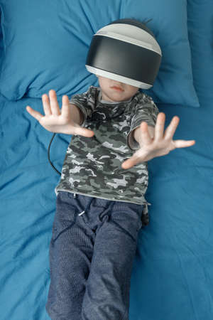 Five-year-old cute boy is lying in his bed in VR glasses playing 3d games. concept of new...