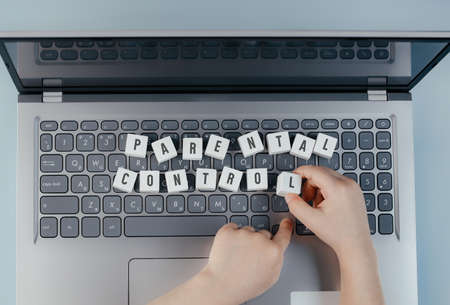 On the keyboard of the laptop there are wooden white cubes with the text Parental control. Childrens hands hold the cube while working at the computer. Child protection conceptの写真素材