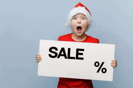 Funny excited white kid in a Santa Christmas costume, screaming and holding a white poster with the...