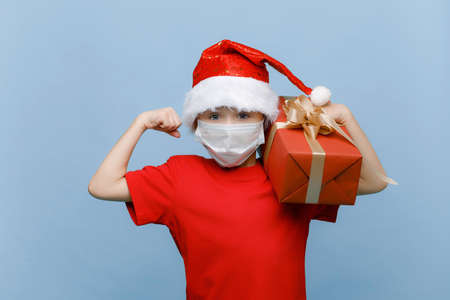 Portrait of a happy little boy in a medical mask, wearing a Christmas hat, holding a gift box and looking into the camera, isolated on a blue backgroundの写真素材