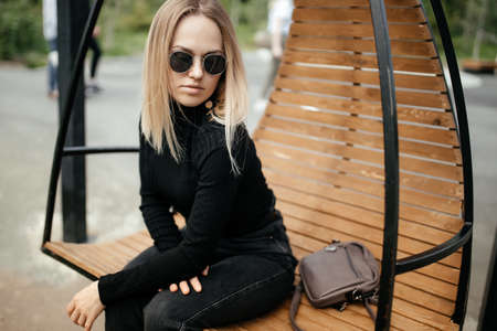 Stylish young blonde is sitting in a round hanging chair outdoor. model sits in a chair and relaxes, looks at the cameraの写真素材