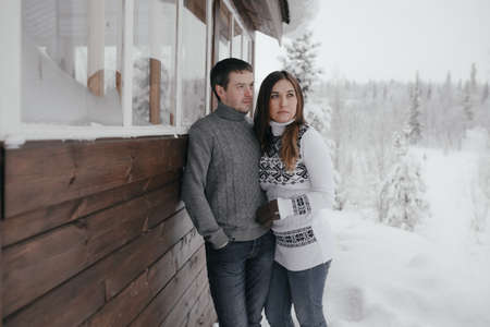 Beautiful couple in love embracing each other, small wooden house and snow in the backgrounの写真素材