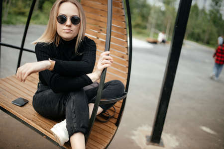 Stylish young blonde is sitting in a round hanging chair outdoor. The model sits in a chair and...
