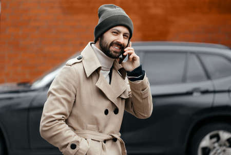 image of a young, smiling, happy, laughing man with a beard in a raincoat with a phone on a gray background on the street. Positive human emotions, facial expression, reaction, attitudeの写真素材