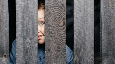 Happy smiling white boy looks out of the crack of a wooden fence. Childish curiosity. Espionage....