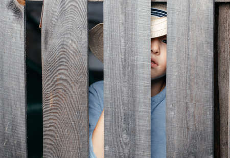 Five- to six-year-old kid in a straw hat peeps through a wooden village fence. Copy spaceの写真素材