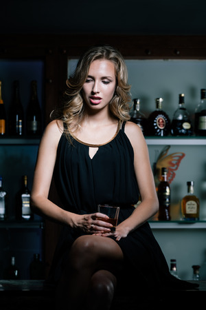 Sad sexy woman sitting on the bar counter with glass of whiskeyの写真素材