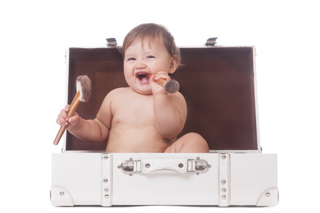 Cheerful little girl with two make-up brushes sitting in a suitcase on isolated whiteの写真素材