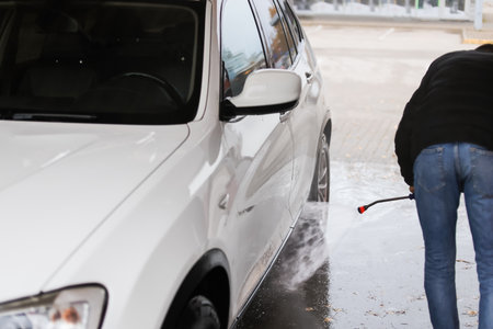 A person rinses a white SUV at a self-service car wash. The scene shows clean water spraying over the vehicle, highlighting the washing process.の写真素材
