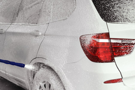 Self-service car wash with foam covering a white SUV. The vehicle is partially cleaned, showing soap suds and water spray from a washing nozzle.の写真素材