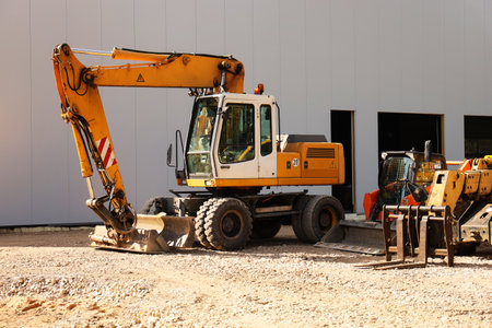 A yellow excavator parked on a construction site. The machine has a large arm and bucket, with gravel scattered around. A building is in the background.の写真素材