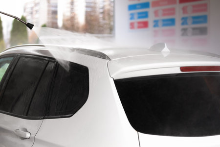 A white car being rinsed with clean water at a self-service car wash. The scene shows a spray nozzle in action, with water droplets visible.の写真素材