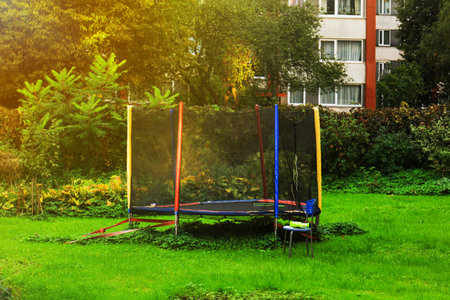 A colorful trampoline sits in a green yard surrounded by trees and shrubs. A residential building is visible in the background.の写真素材