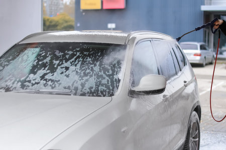 A person rinses a silver SUV with clean water at a self-service car wash. Soap suds cover the vehicle's surface, and a modern wash station is visible in the background.の写真素材