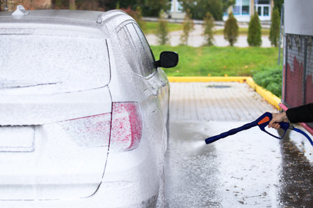 Self-service car wash with foam covering a white vehicle. A person is using a pressure washer to clean the car. The setting is outdoors with greenery in the background.の写真素材