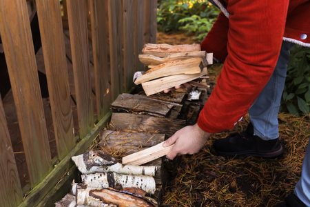 The man with short brown hair stacks firewood beside a wooden fence. The scene is set in a garden with green foliage and fallen leaves.の写真素材