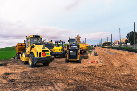 Road construction scene with a road roller, excavator, mini dump truck, and tractor on a dirt road. Heavy machinery is parked on the site with a cloudy sky in the background.の写真素材