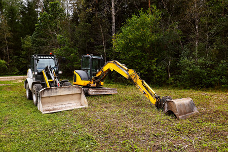 A crawler excavator and a tractor are parked in a forest clearing. The area is surrounded by trees and greenery, showing a natural environment.の写真素材