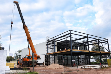 A truck crane operates at a construction site with a new building structure. The area is surrounded by fencing and materials are scattered around.の写真素材