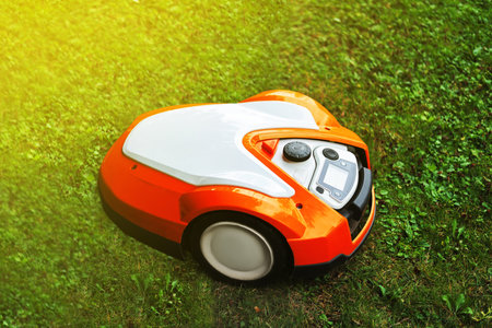 A robotic lawn mower sits on a green lawn. The mower is orange and white, designed for automated grass cutting. Sunlight illuminates the scene.の写真素材