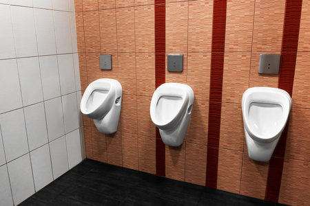 Three white urinals in a public restroom with tiled walls and dark floor. High quality photoの写真素材