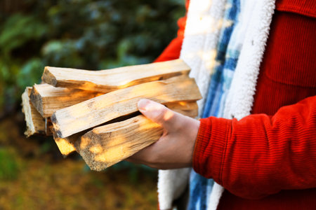 The man holds a bundle of firewood in a forest setting. He wears a red jacket and a plaid shirt, surrounded by greenery.の写真素材