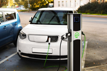 A white electric car is charging at a designated parking lot for electric vehicles. A charging station is visible next to the car, with green cables connected.の写真素材
