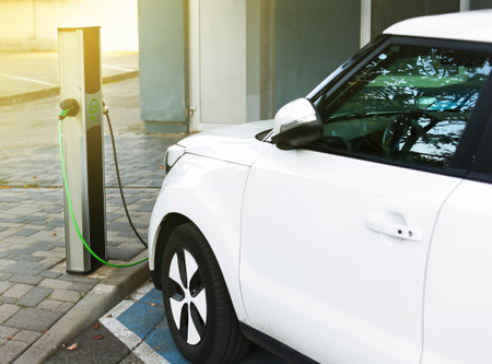 A white electric car is charging at a designated parking spot in an electric vehicle charging station. The setting is a modern urban environment with sunlight.の写真素材