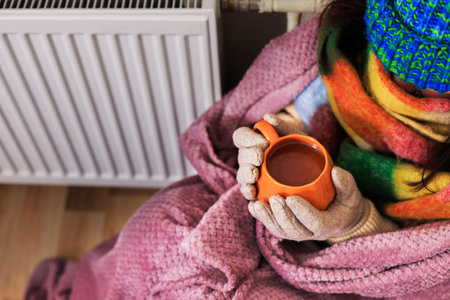A close-up of a girl holding a cup of tea. She sits near a radiator. She's wearing a scarf, gloves, and a sweater. She's wrapped in a purple blanket. She's trying to keep warm in a poorly heated room,の写真素材